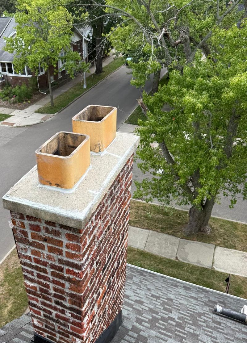 Red brick chimney with two clay flue tiles and a concrete crown above a tree-lined street